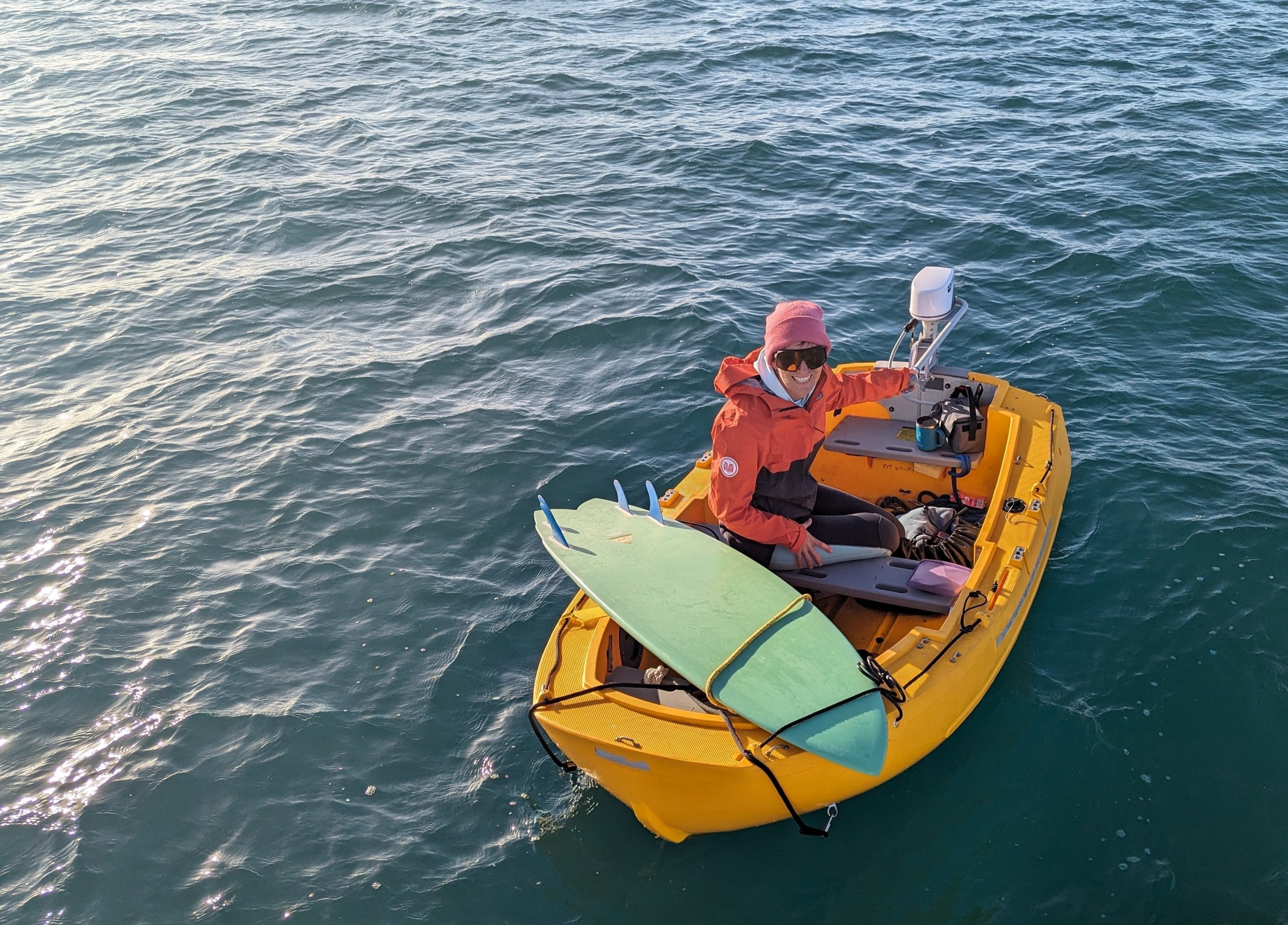 Woman in a wetsuit in a small yellow boat with a green surfboard strapped on top.