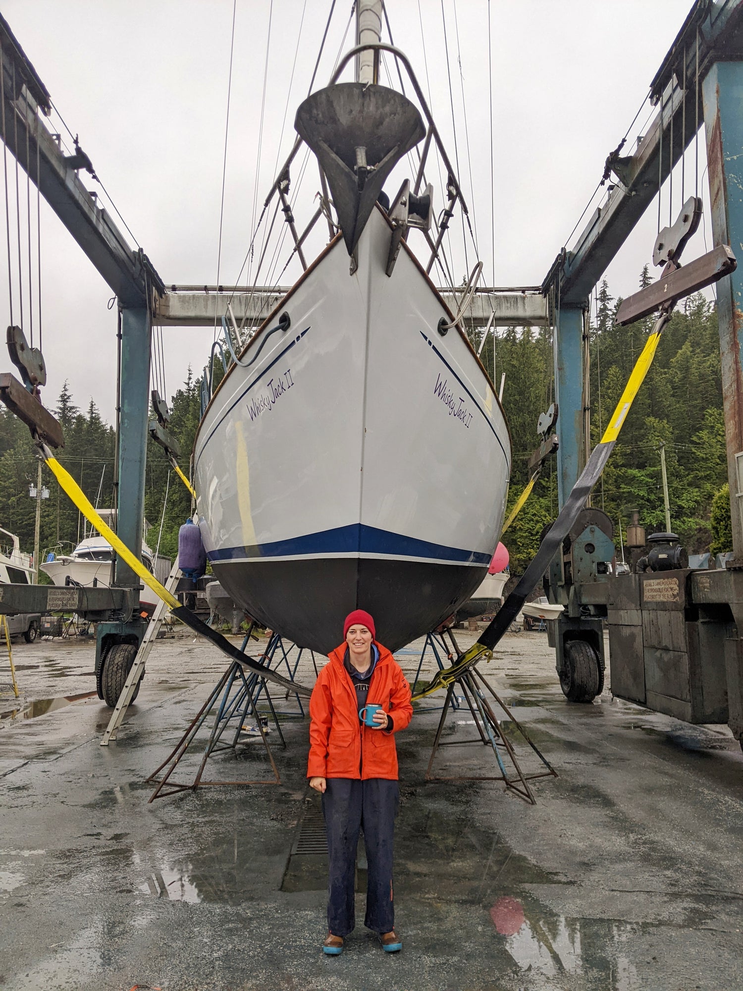 The bow of a Kelly Peterson 44 sailboat, in slings on the hard, with a persone in an orange raincoat standing in front of it.